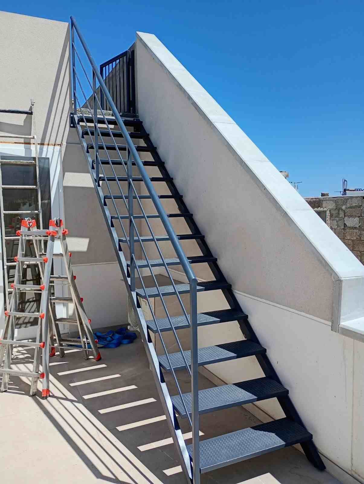 Escalera metálica exterior en una azotea junto a paredes blancas bajo un cielo azul despejado.