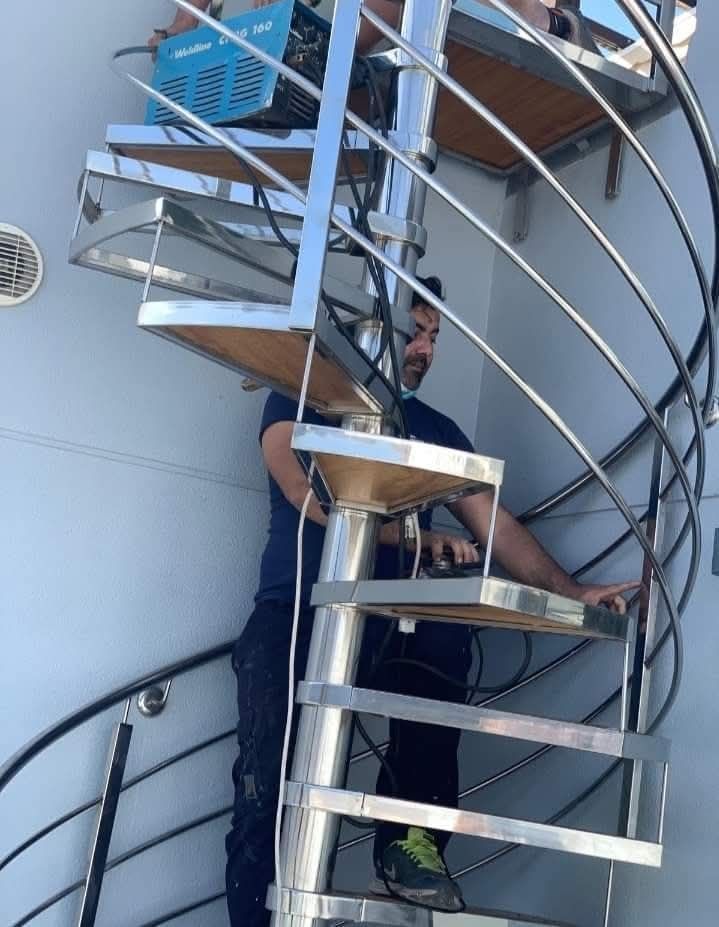 Un hombre sentado en una escalera de caracol al aire libre, sosteniendo un palo entre escalones blancos y azules.