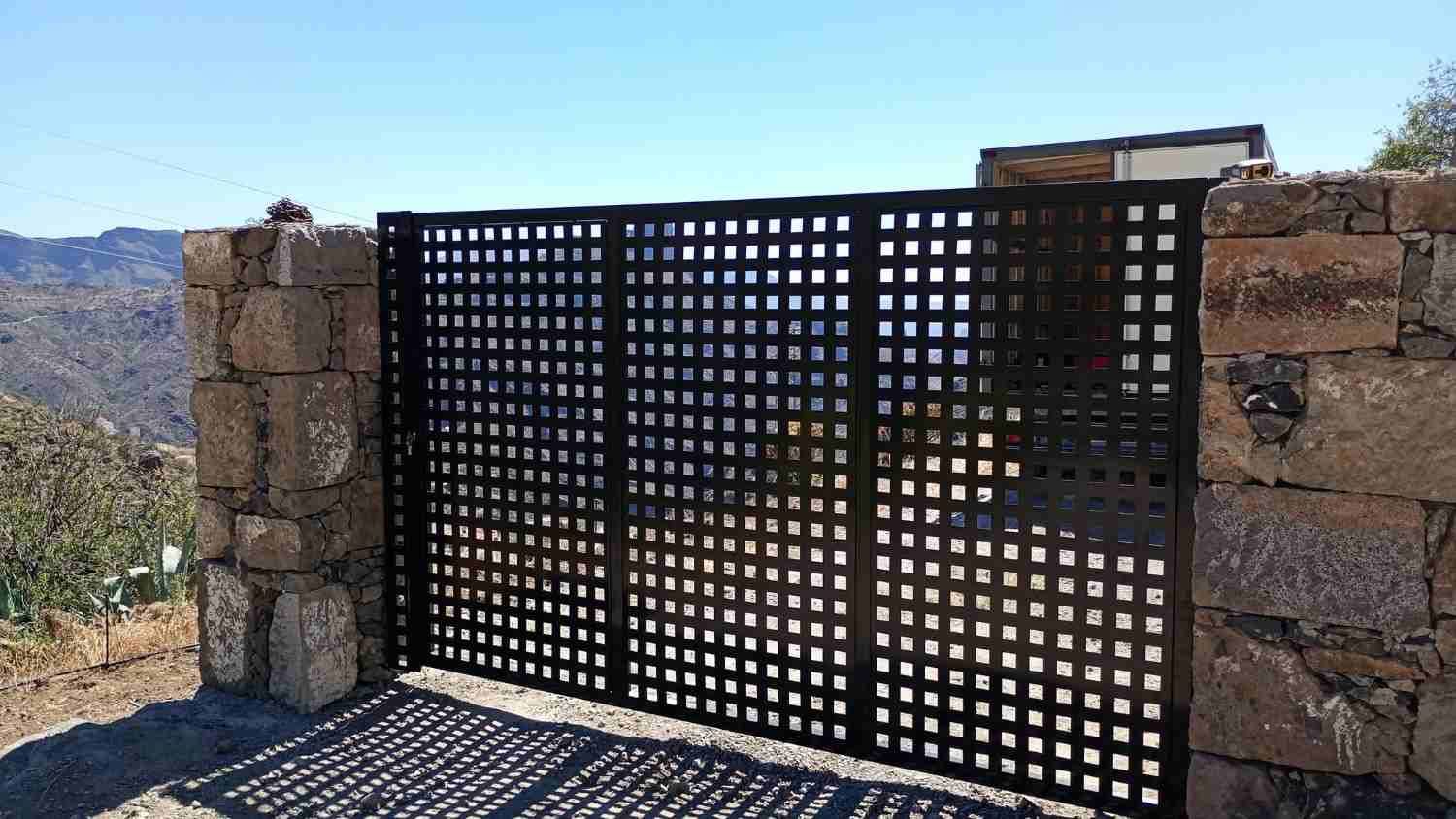 Puerta de metal negro entre pilares de piedra con vistas a una soleada montaña.