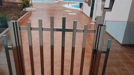 Una verja metálica bloquea un camino de baldosas mojadas junto a un edificio y un sendero ajardinado.