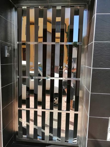 Puerta metálica vertical con listones estrechos en un marco negro, que deja ver un atisbo del interior que hay detrás.