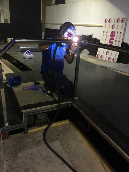 Una persona suelda sobre una plataforma metálica en un taller con poca luz; las chispas brillantes iluminan la escena.