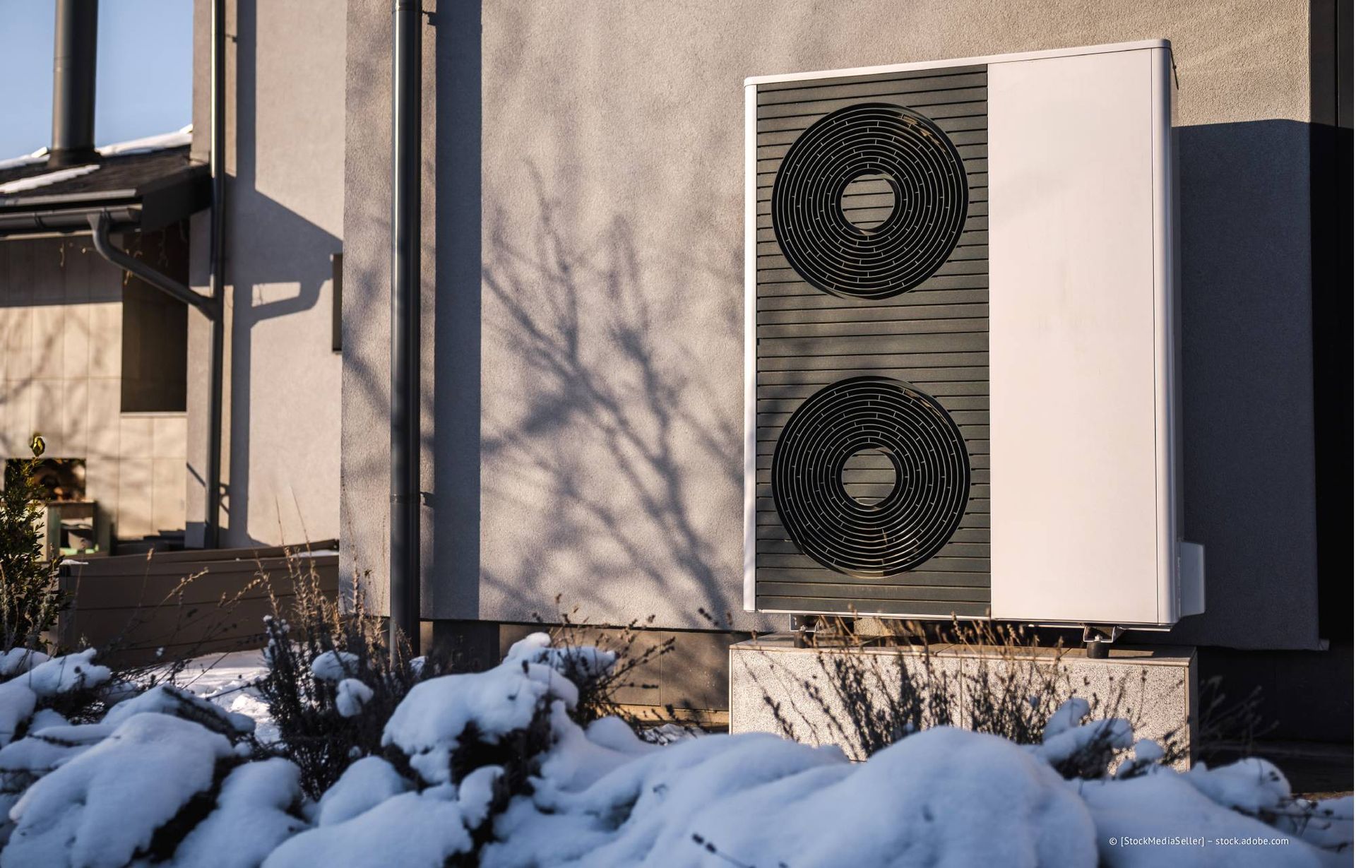 Wärmepumpeneinheit außerhalb eines Hauses, schneebedeckter Boden und Wintersonne.