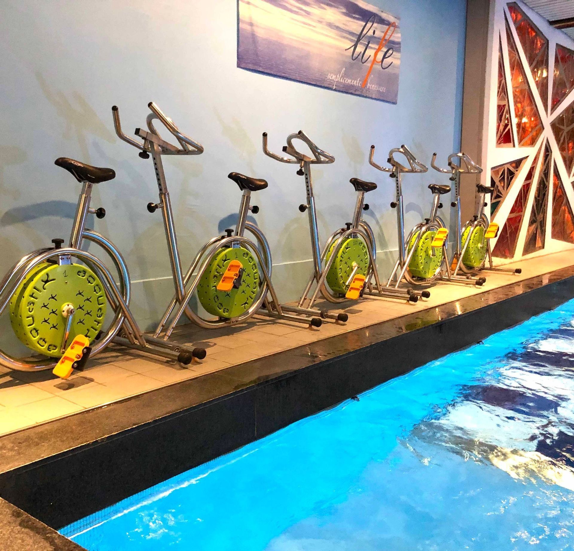 Cyclette allineate in una piscina poco profonda, acqua turchese, accenti gialli, luce del sole.