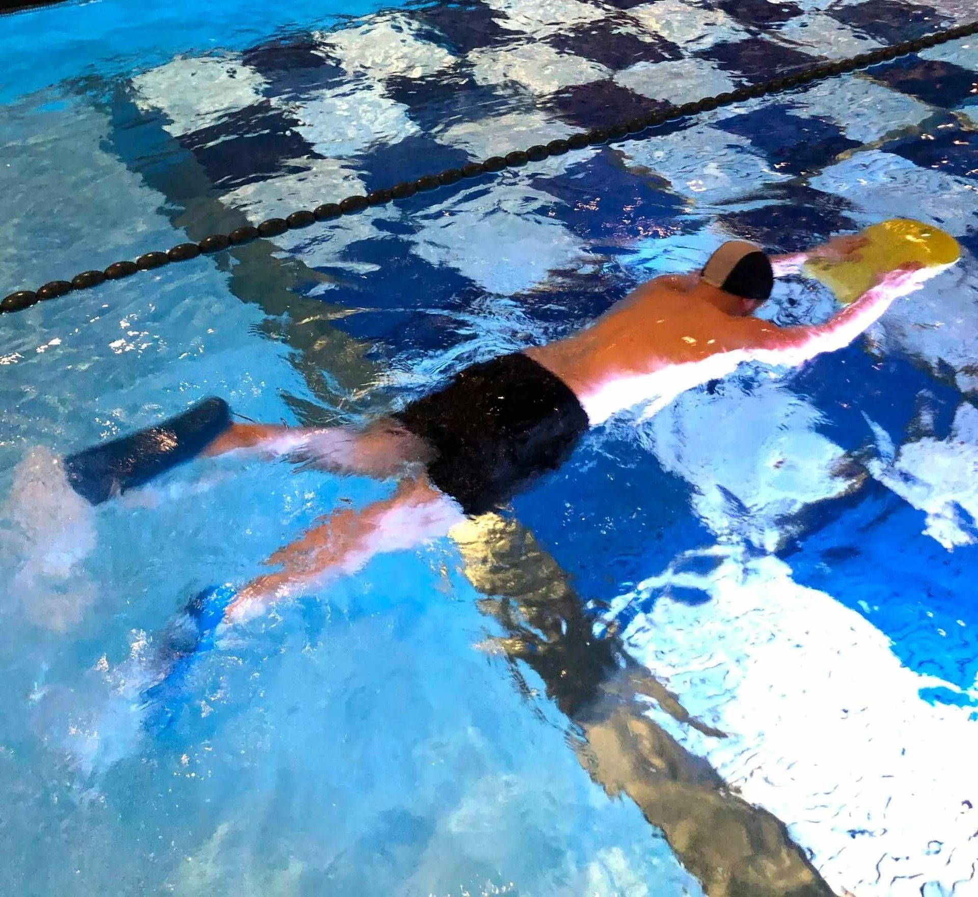 Nuotatore in piscina, con in mano una tavoletta gialla, indossa pantaloncini neri e pinne.