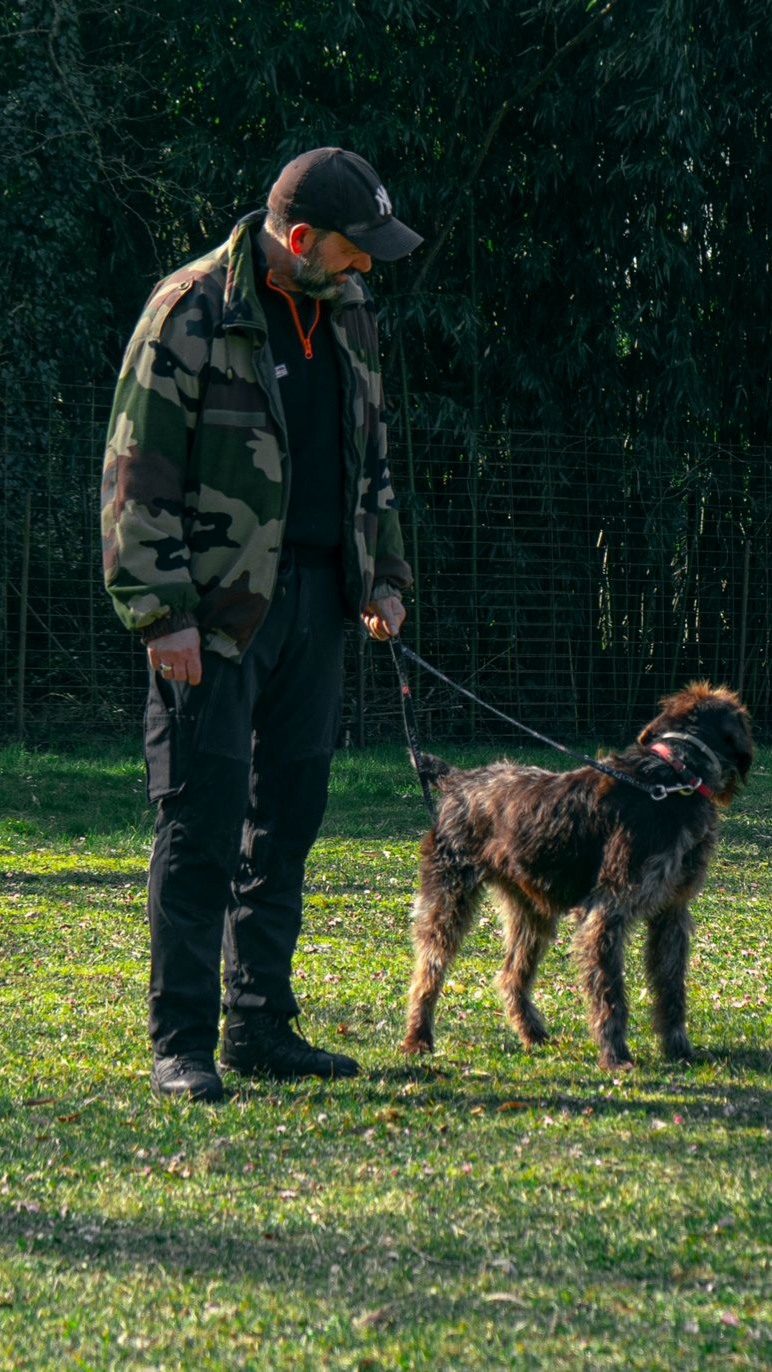 Un homme en veste de camouflage tient un chien en laisse dans une zone herbeuse.