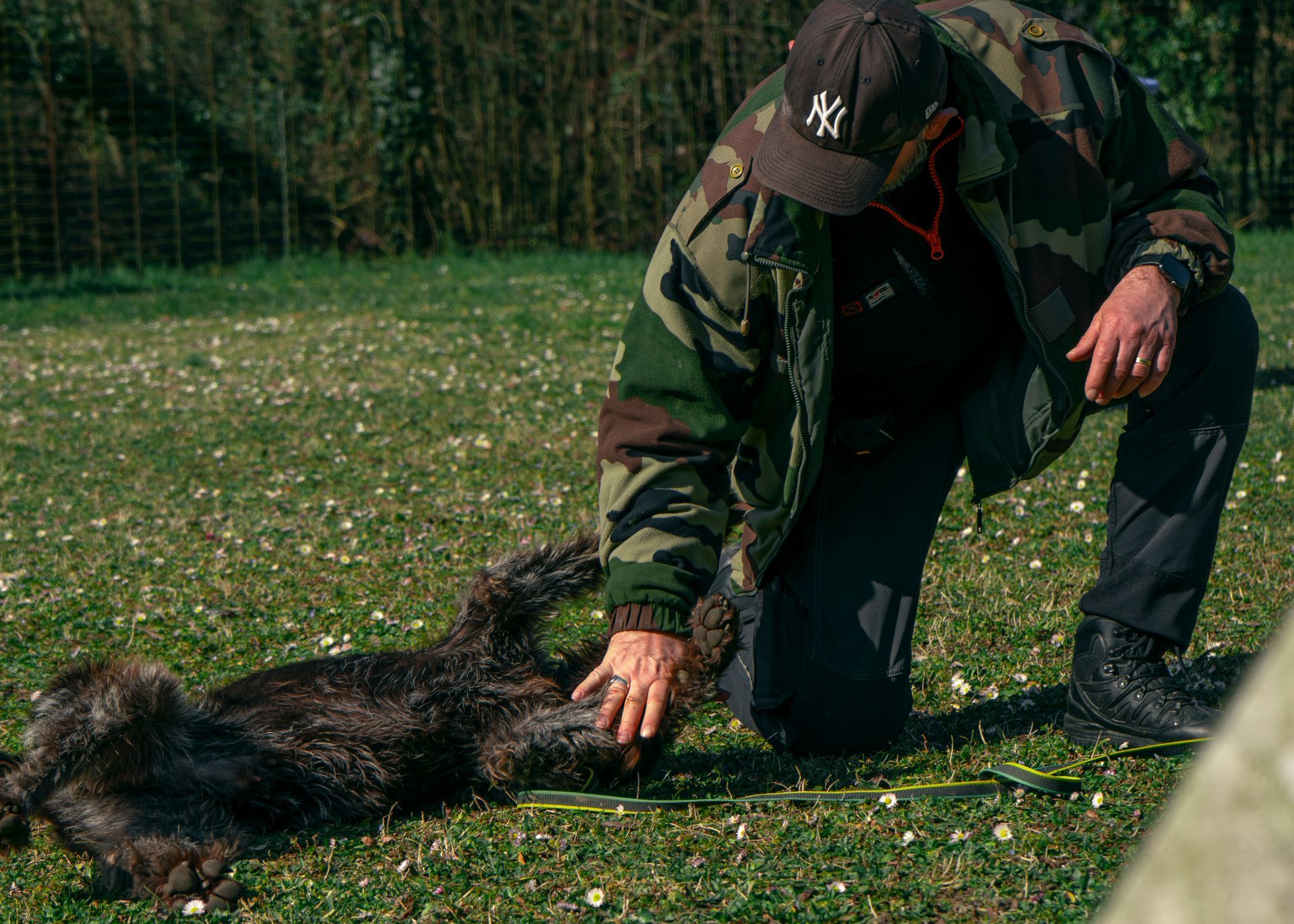 Une personne en tenue de camouflage est agenouillée et caresse un chien au pelage sombre.
