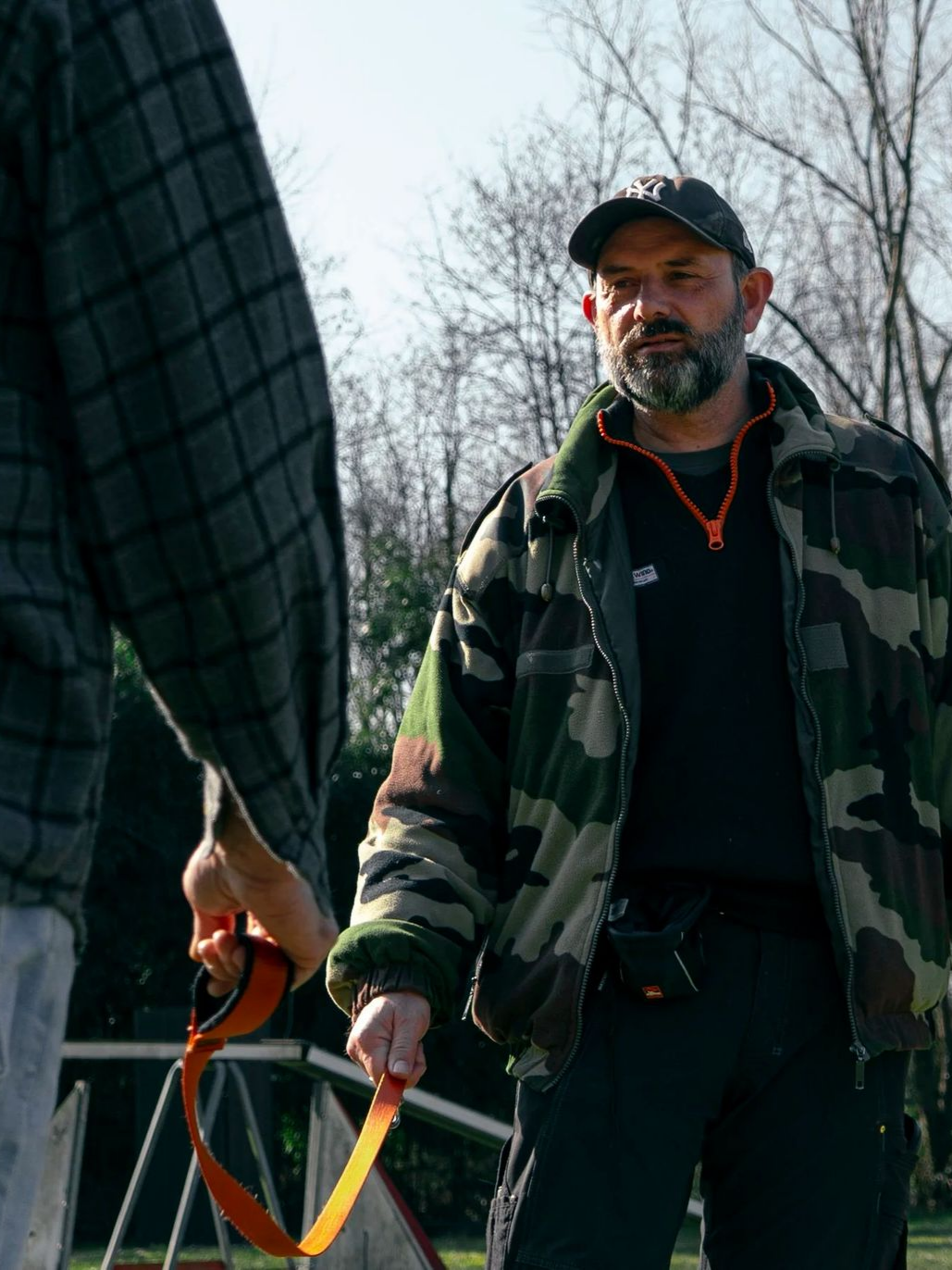 En extérieur, un homme en veste de camouflage tient une laisse de chien et regarde une autre personne.