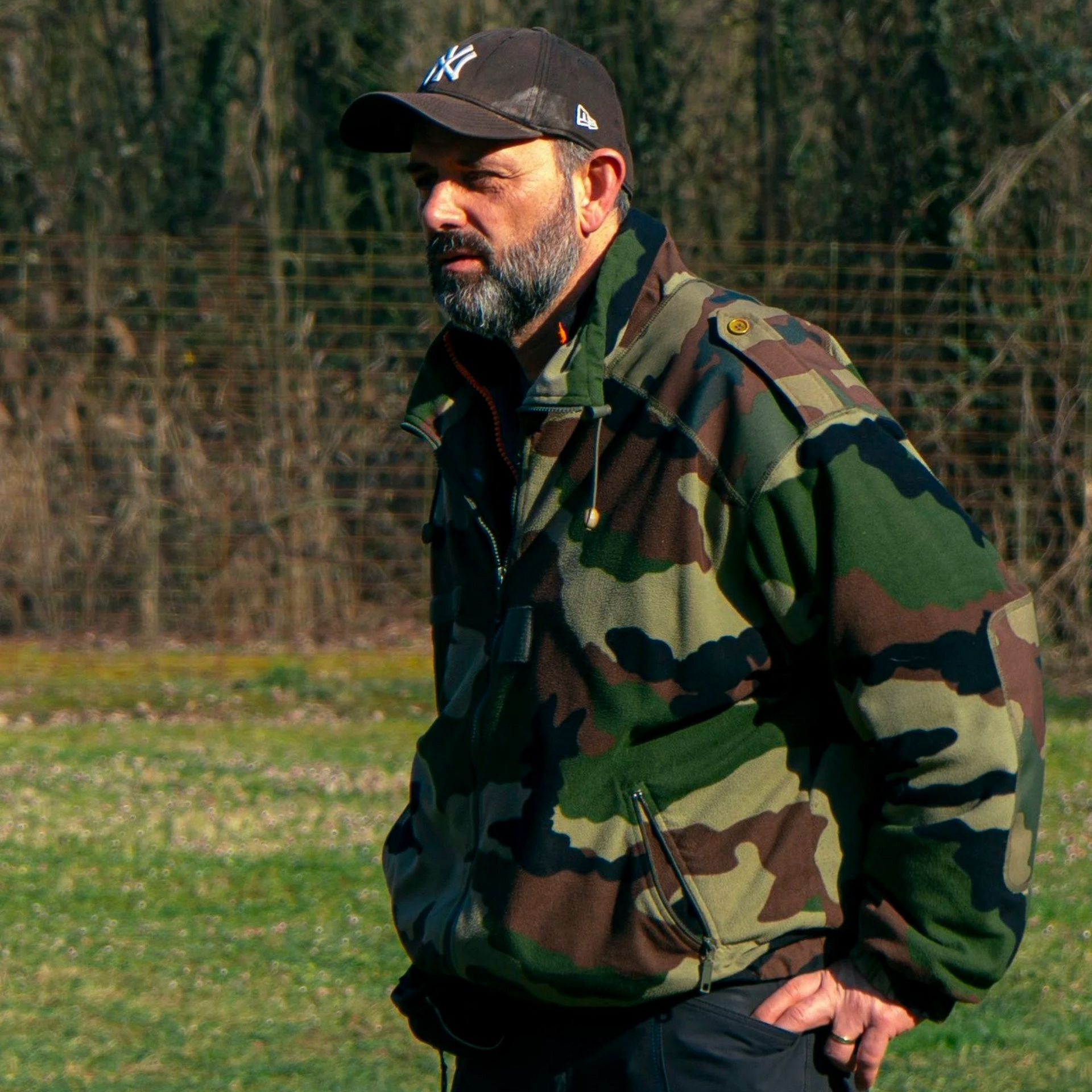 Un homme portant une veste de camouflage et une casquette se tient debout à l'extérieur.