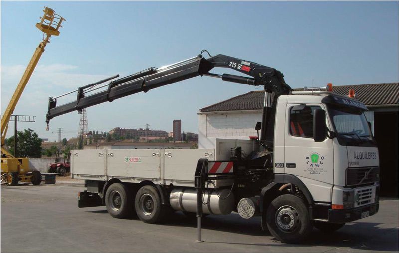Un camión blanco de plataforma con una gran grúa negra montada detrás de la cabina, estacionado en un aparcamiento asfaltado.