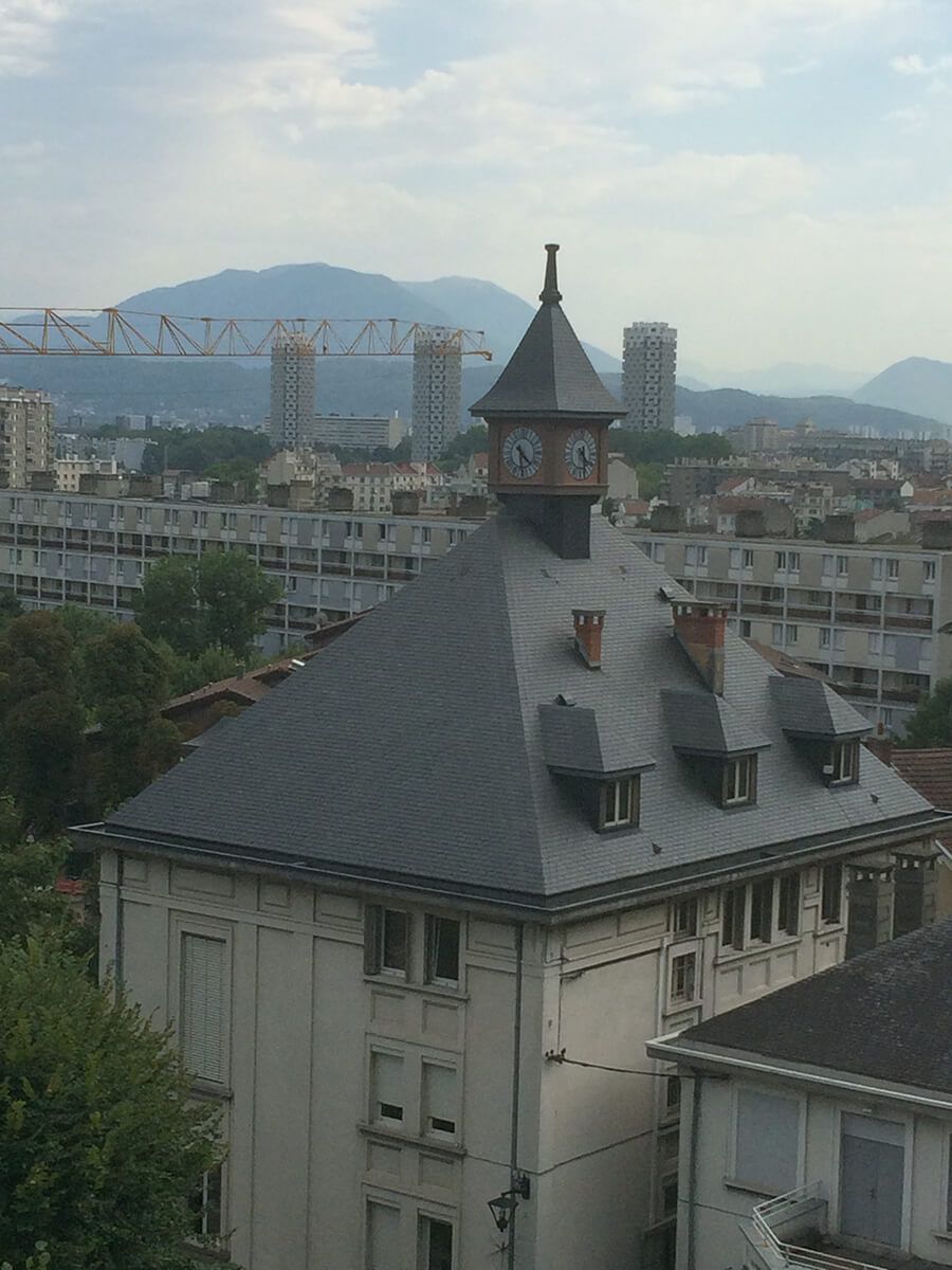 Bâtiment historique avec un toit en ardoise pentu et une tour, surplombant une ville avec des montagnes au loin