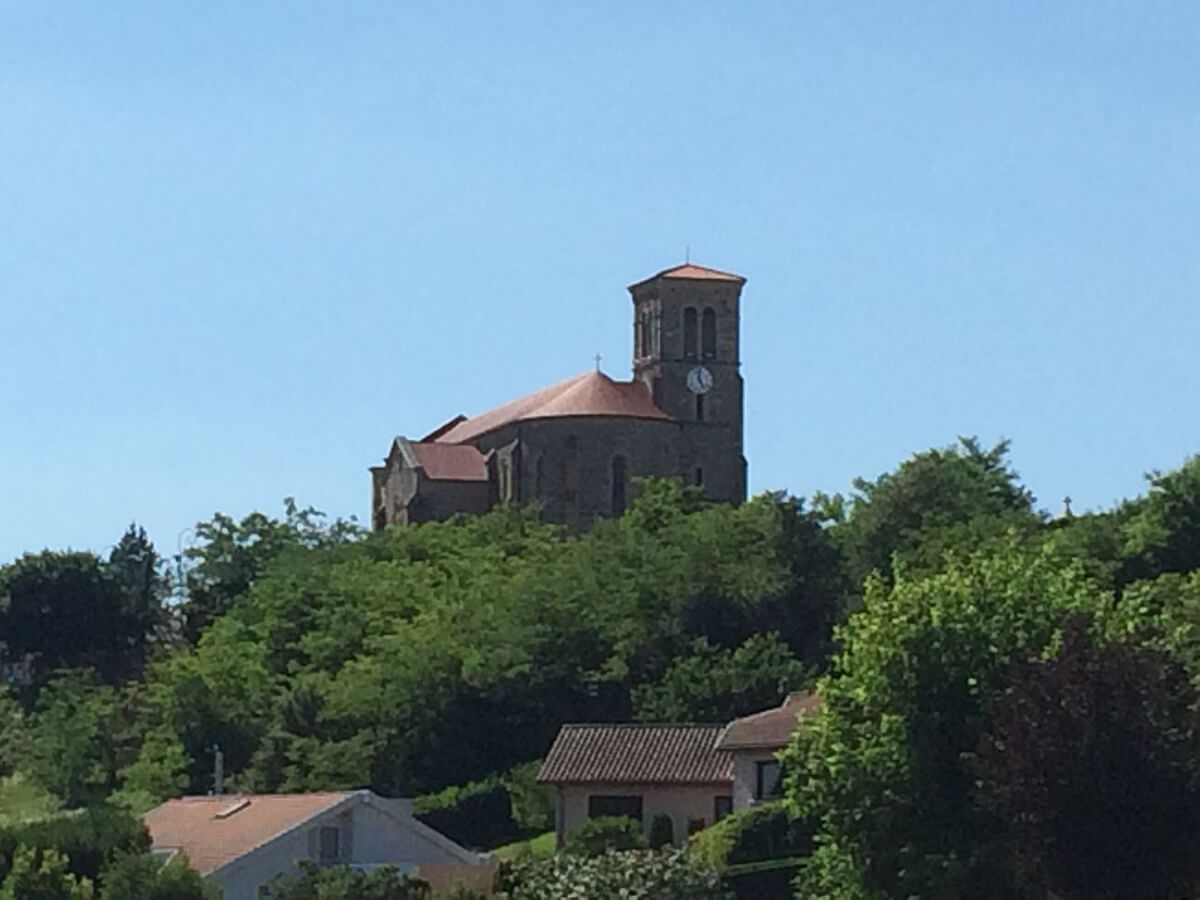 Église perchée sur une colline, au toit rouge, surplombant des arbres verdoyants et des maisons sous un ciel bleu dégagé