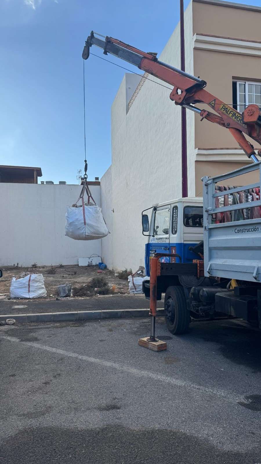 Una grúa levanta una bolsa de escombros de un camión junto a la pared de un edificio en un estacionamiento.
