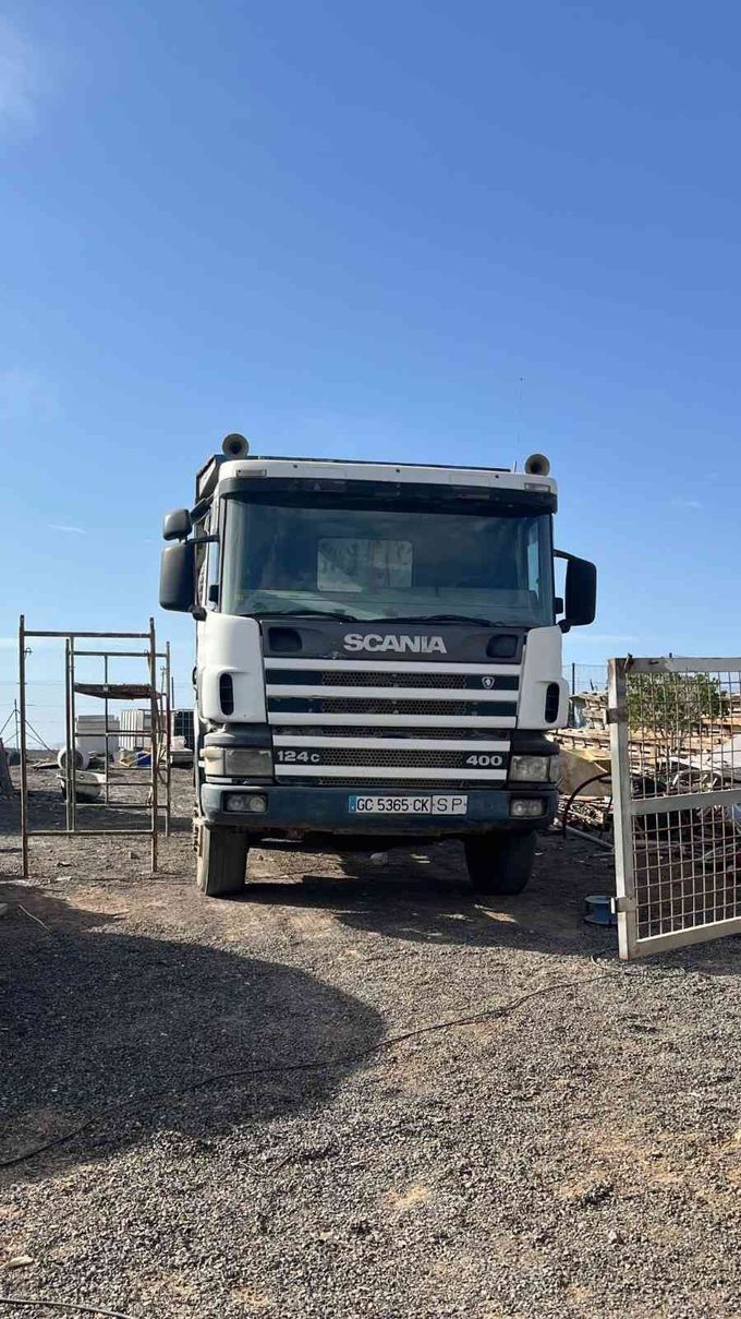 Camión Scania blanco estacionado sobre grava dentro de un patio industrial cercado bajo un cielo azul despejado.