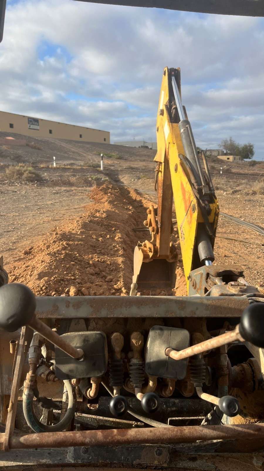 Brazo de excavadora removiendo tierra en una obra de construcción bajo un cielo parcialmente nublado.