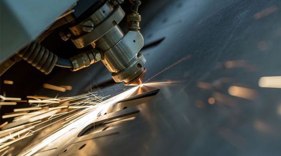 Primer plano de un cabezal de corte láser fundiendo una lámina de metal, creando chispas de color naranja brillante y material fundido.