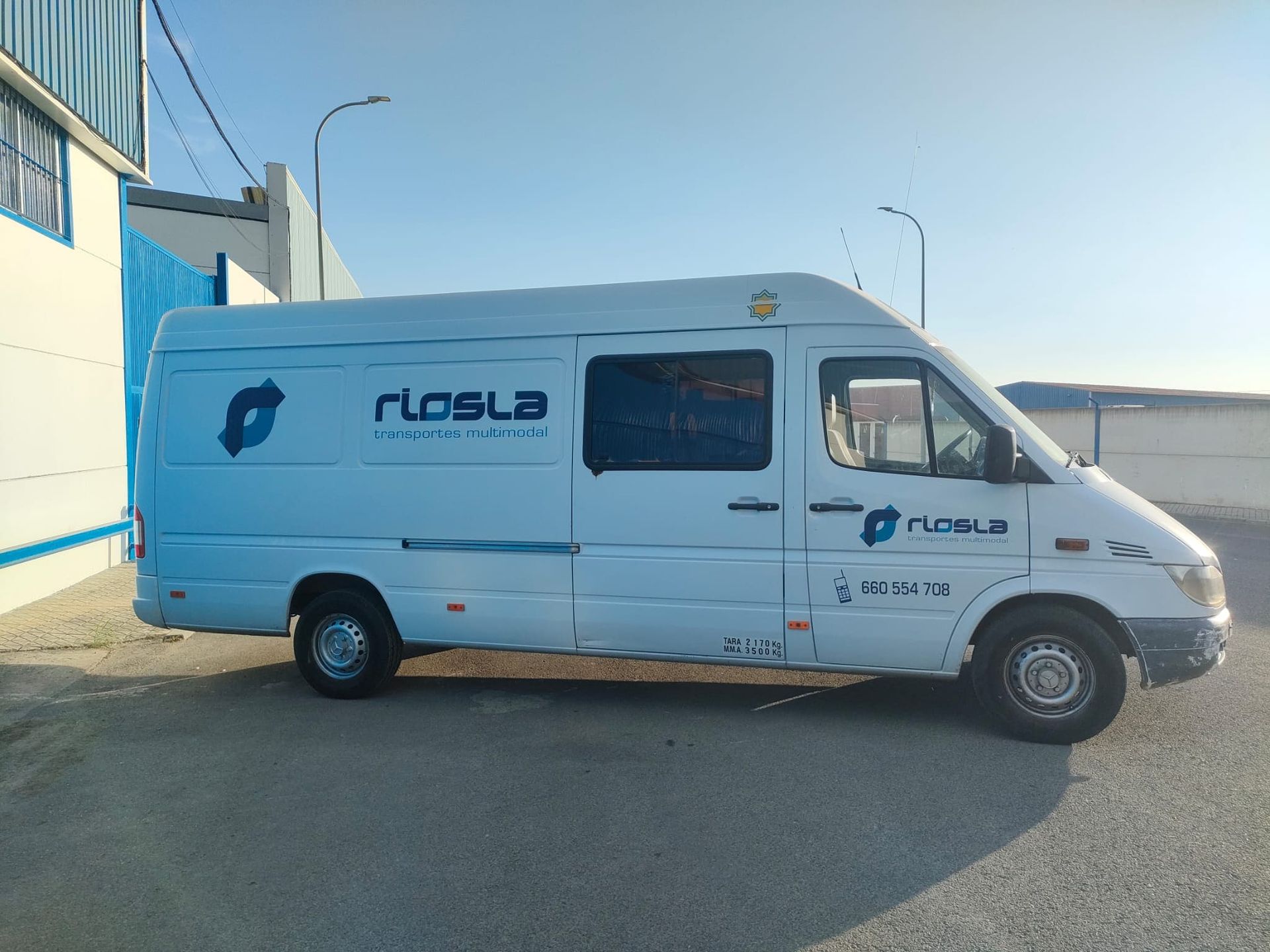 Furgoneta blanca Riosla estacionada cerca de un edificio azul bajo un cielo despejado.