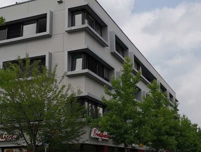 Graues Gebäude mit dunkel gerahmten Fenstern und grünen Bäumen davor. „Pizzeria“-Schild sichtbar.