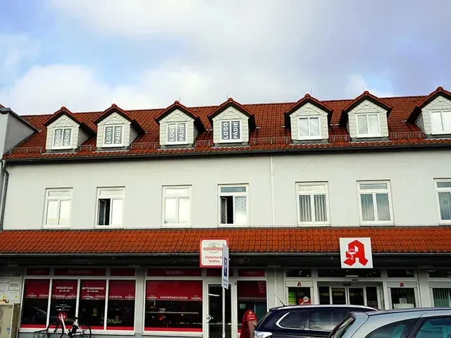 Außenansicht eines weißen Gebäudes mit roten Dachziegeln und Dachgauben, im Erdgeschoss befindet sich eine Apotheke.