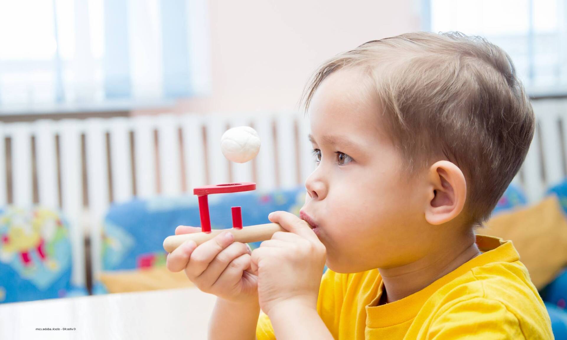 Kleines Kind bläst Luft in einen Ball auf einem kleinen Tisch. Drinnen mit einem weißen Tisch und blauen Möbeln.