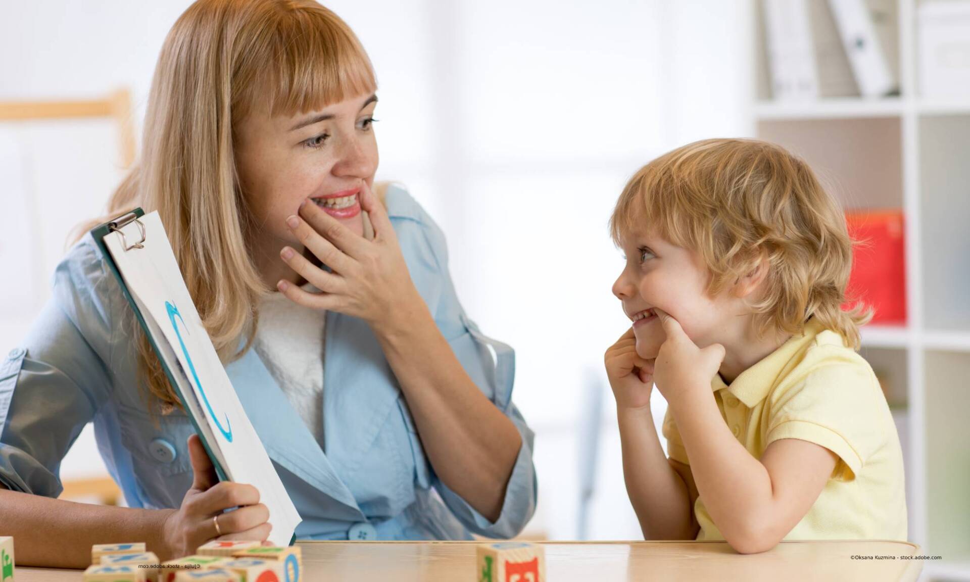 Frau mit Klemmbrett und lächelnder Junge an einem Tisch in einem Raum.