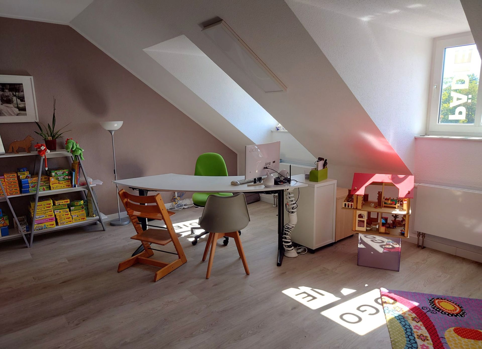 Büroraum mit schräger Decke und weißem Schreibtisch, zwei grünen Stühlen, einem orangefarbenen Stuhl. Regale mit Spielzeug.