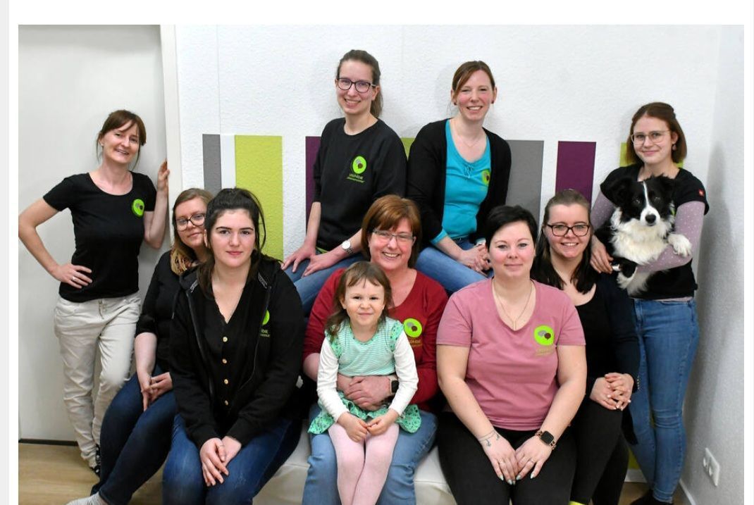 Eine Gruppe von Frauen und ein Kind lächeln in die Kamera, einige haben die Arme umeinander gelegt, vor farbenfrohen Wandakzenten.