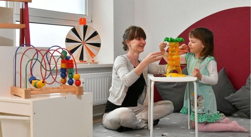 Frau und Kind spielen mit Spielzeug in einem hell erleuchteten Spielzimmer; die Frau lächelt und interagiert mit dem Kind.