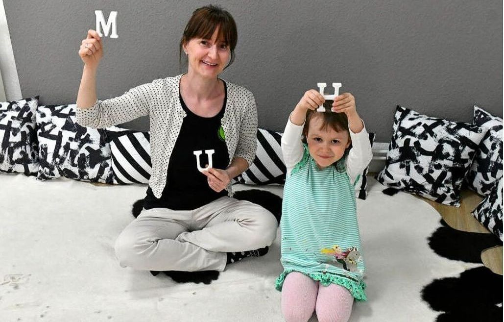 Frau und Kind halten die Buchstaben M und U; sitzen auf dem Boden mit schwarzen und weißen Kissen.