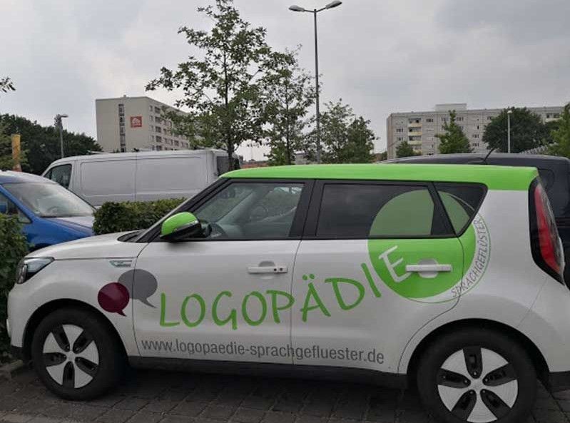 Weißes Auto mit grünen Akzenten, „LOGOPÄDIE“-Logo, vor Gebäuden geparkt.