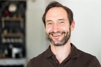 Ein Mann mit Bart und einem braunen Hemd lächelt in die Kamera.