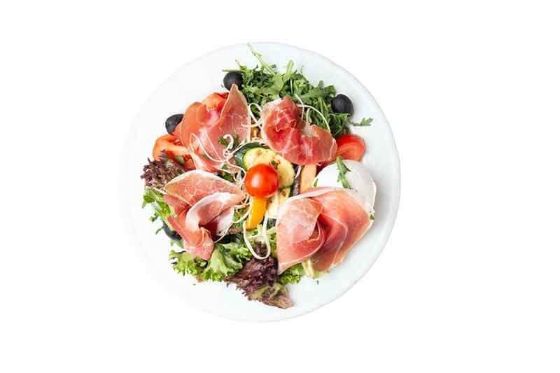 Salat mit Prosciutto, Tomaten, Oliven und Mozzarella auf einem weißen Teller.