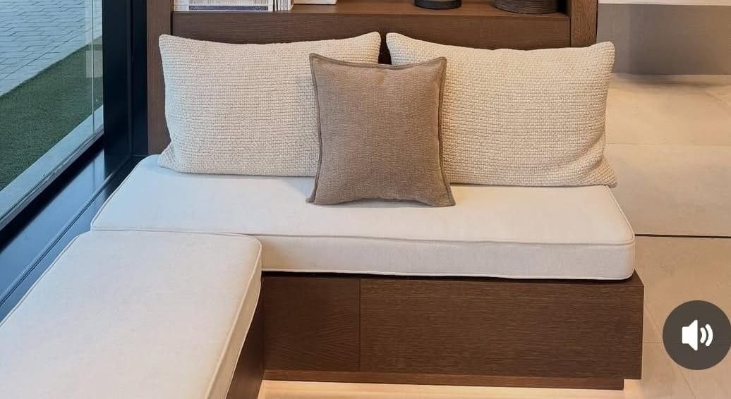 Un banco empotrado de color marrón con cojines blancos, dos almohadas texturizadas de color claro y un cojín decorativo cuadrado de color marrón liso.