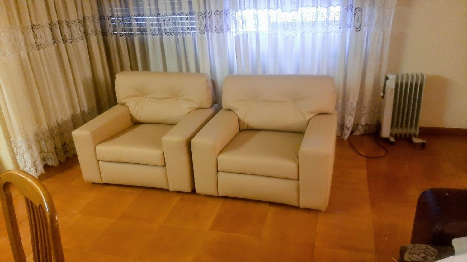 Dos sillones de cuero beige colocados uno al lado del otro frente a unas cortinas blancas, con un radiador portátil cerca.