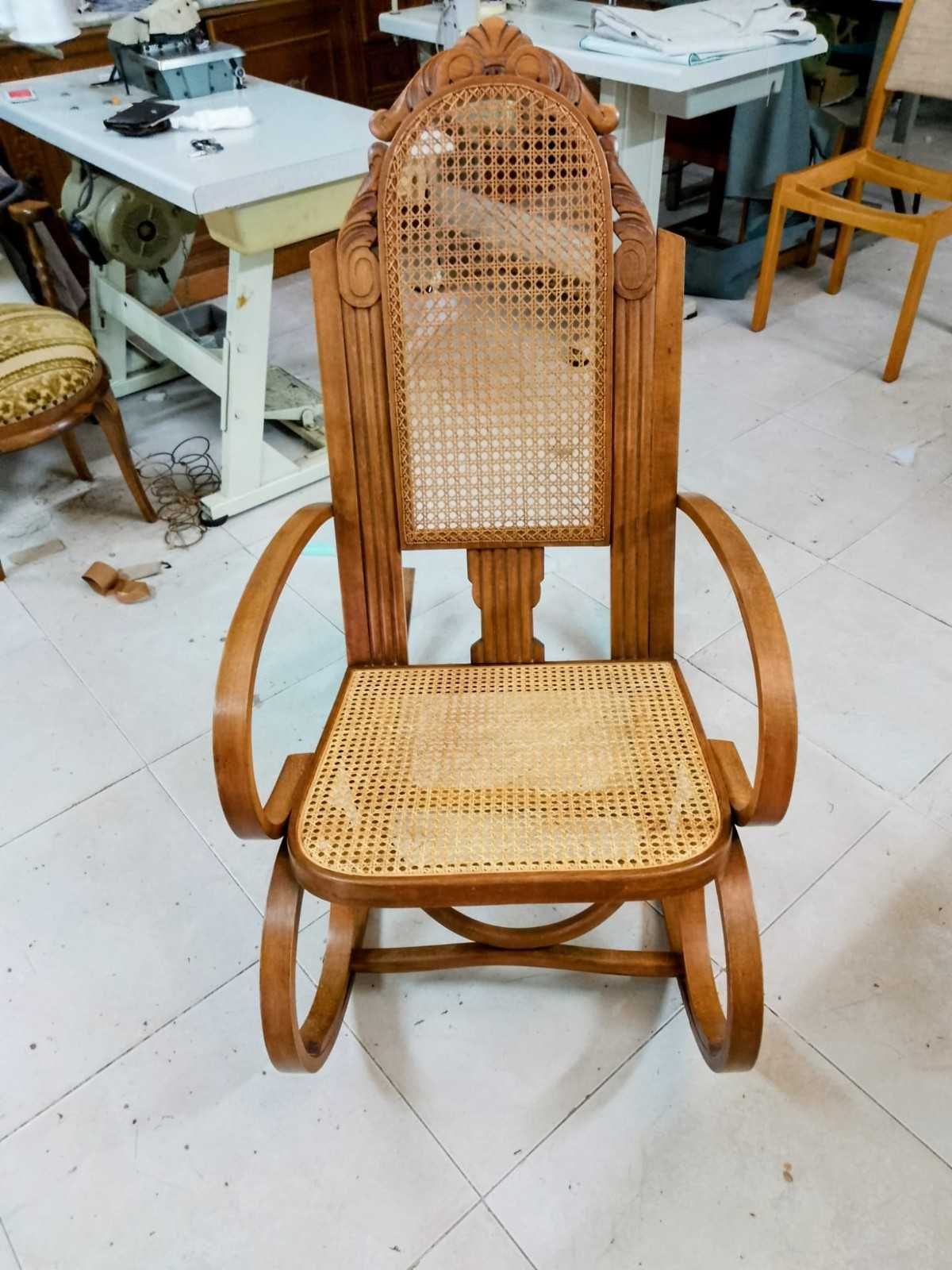 Una mecedora de madera con asiento y respaldo de mimbre, situada en una habitación con mesas de taller y máquinas de coser.