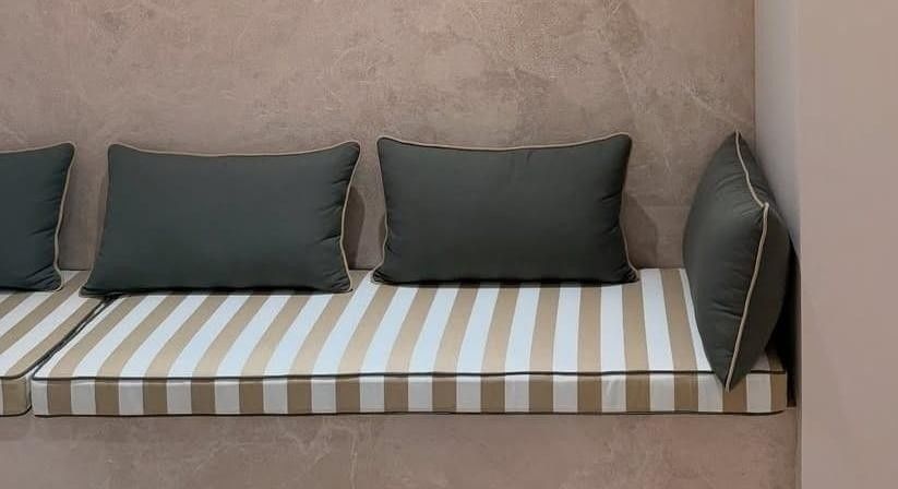 Un banco con un cojín a rayas blancas y beige, tres almohadas de color carbón y una pared de fondo texturizada de color rosa pálido.