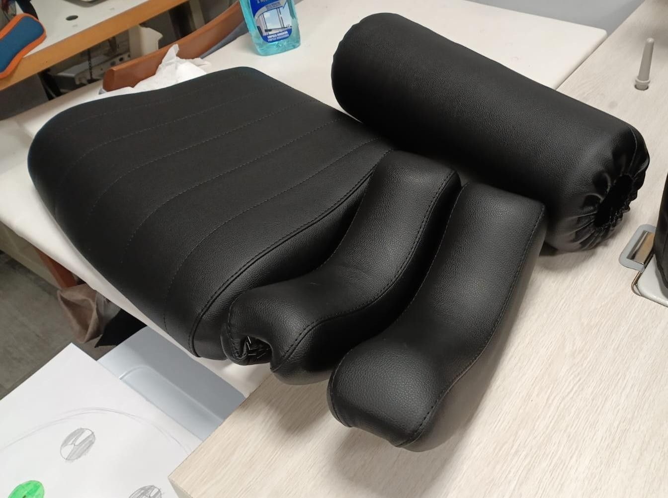 Sobre un banco de trabajo de color claro, se encuentran piezas tapizadas en cuero negro, que incluyen un asiento, dos reposabrazos y un almohadón.