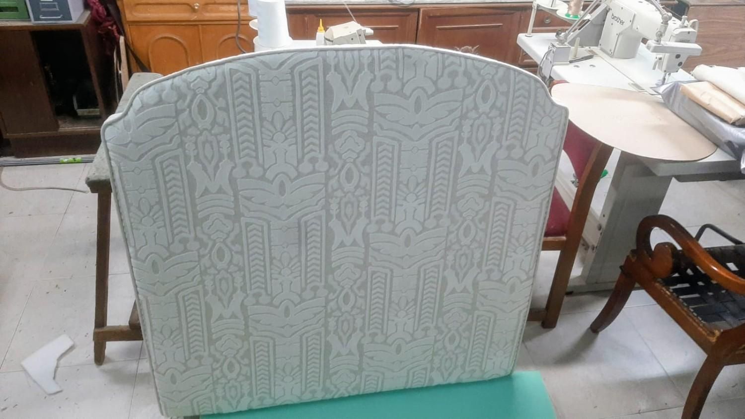 En un taller, junto a una máquina de coser, se encuentra el respaldo de una silla retapizado con una tela estampada de color claro.