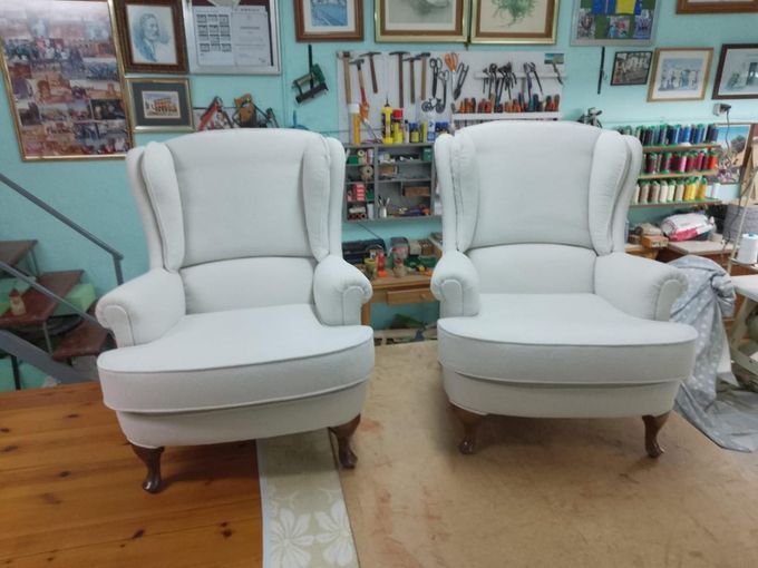 Dos sillones orejeros blancos, recién tapizados, se encuentran uno al lado del otro en un taller de muebles bien equipado.