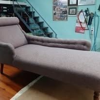 Una tumbona capitoné de color lila claro descansa sobre un suelo de madera en una habitación con paredes azul claro y una escalera metálica.