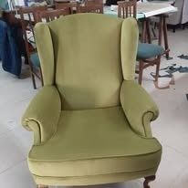 En una habitación, al fondo, se ve un sillón orejero de terciopelo verde lima, con otras sillas y mesas de madera.