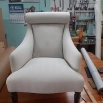 Un sillón tapizado de color gris claro, con respaldo redondeado y patas de madera, se encuentra en un taller artesanal.