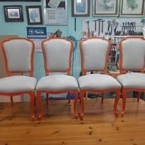 En un taller, cuatro sillas de comedor tapizadas en gris claro con estructura de madera están alineadas sobre una mesa de madera.