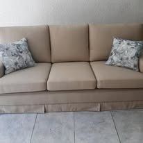 Un sofá beige de tres plazas con faldón de tela y dos cojines decorativos estampados, sobre un suelo de baldosas.