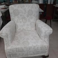 Un sillón de color blanco roto con un delicado estampado floral repetitivo, situado en una habitación con un taburete al fondo.