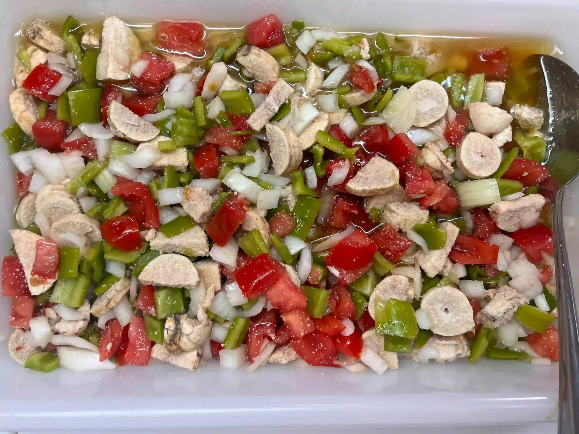 Verduras rojas y verdes picadas mezcladas con trozos blancos en rodajas en una bandeja transparente, cubiertas con aderezo.