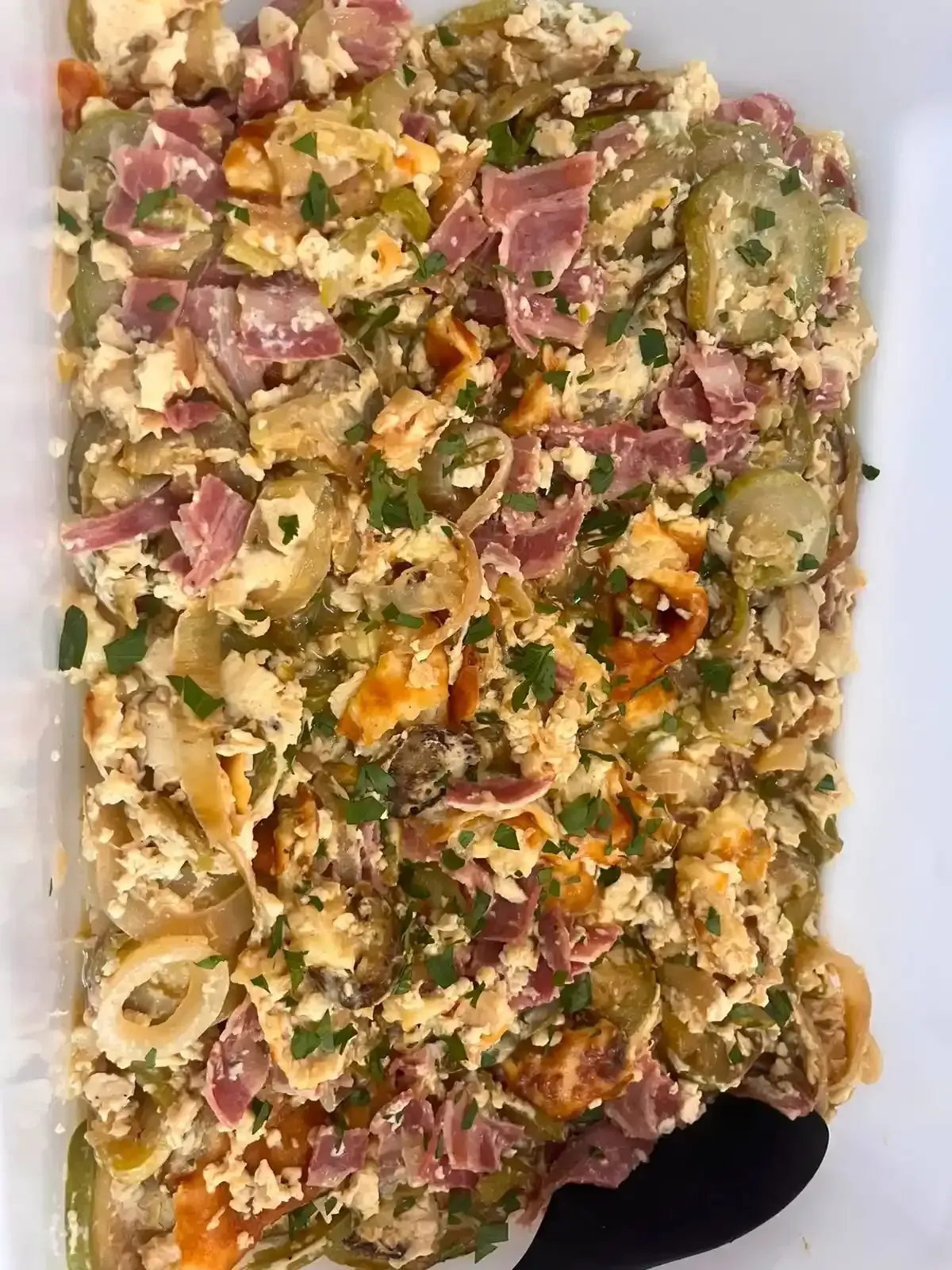 Bandeja de ensalada de pasta mixta con jamón, queso y hierbas, coronada con una cuchara de servir negra.