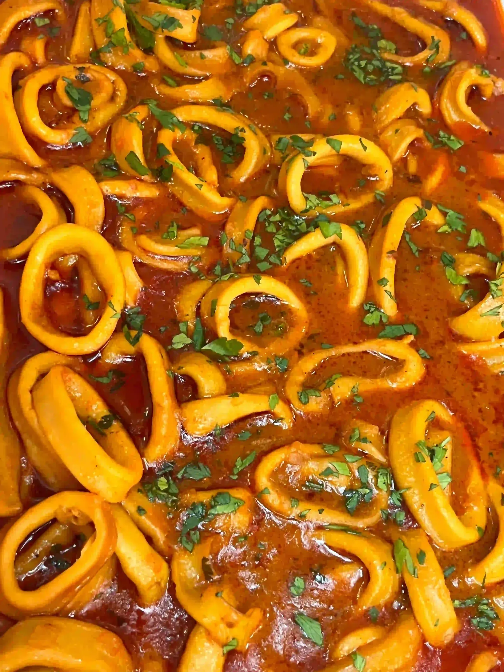 Primer plano de aros de calamar cociéndose a fuego lento en una rica salsa de color naranja rojizo con hierbas picadas.