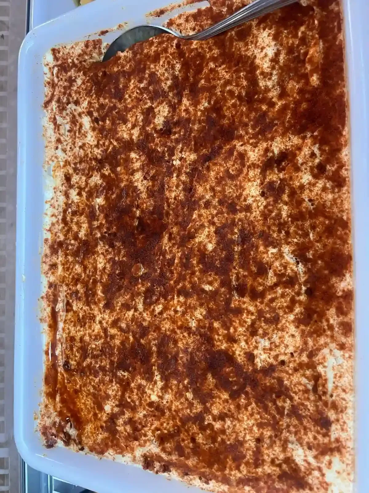 Postre rectangular en un plato transparente, cubierto con cacao en polvo, con una cuchara en el borde superior.