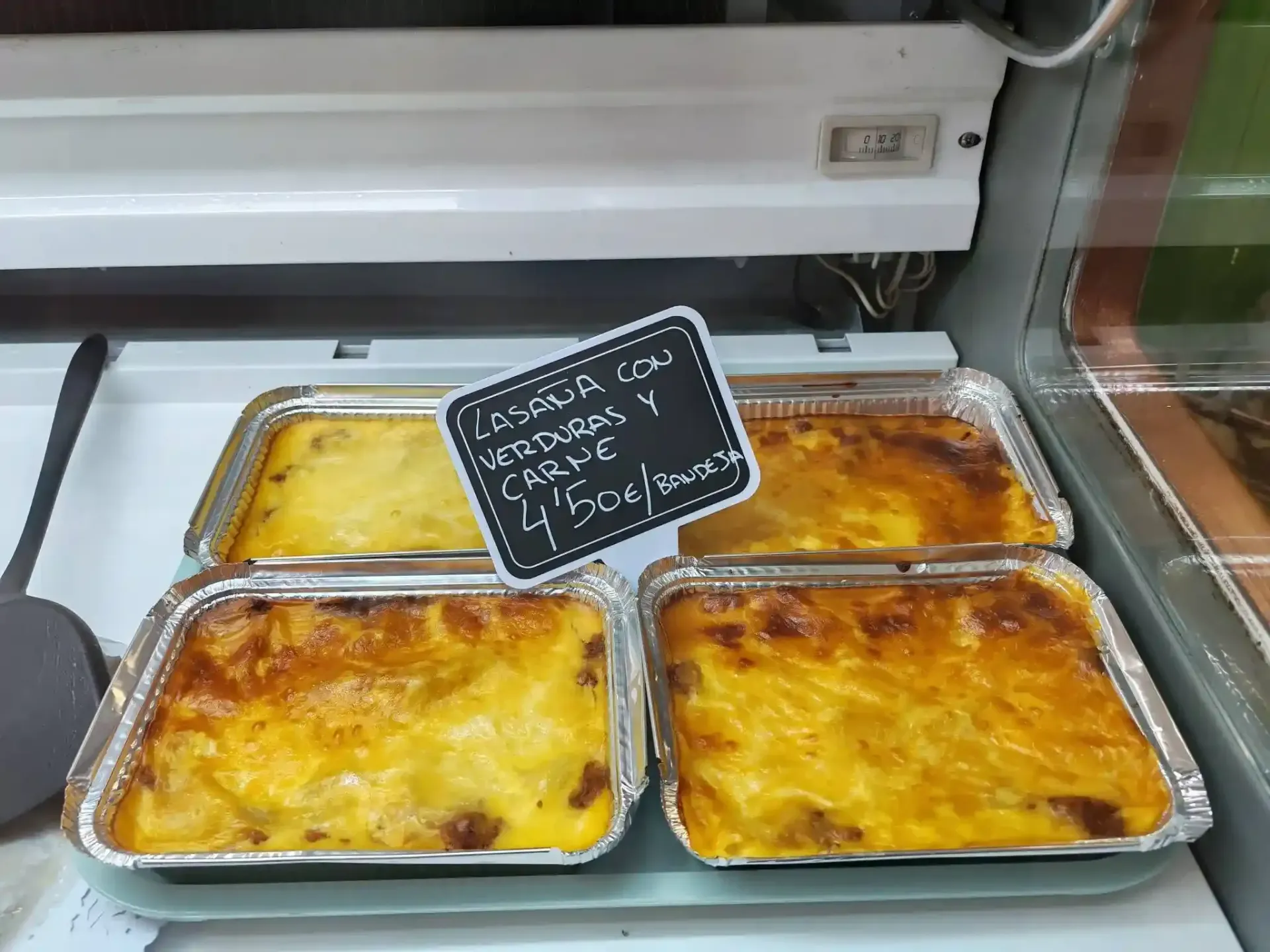 Bandeja de guisos horneados en una vitrina con un cartel de precio escrito a mano.